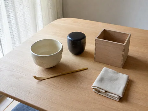 日本の住まいの低い机に置かれた茶碗、茶杓、棗、無地の木箱と包み布のイメージ